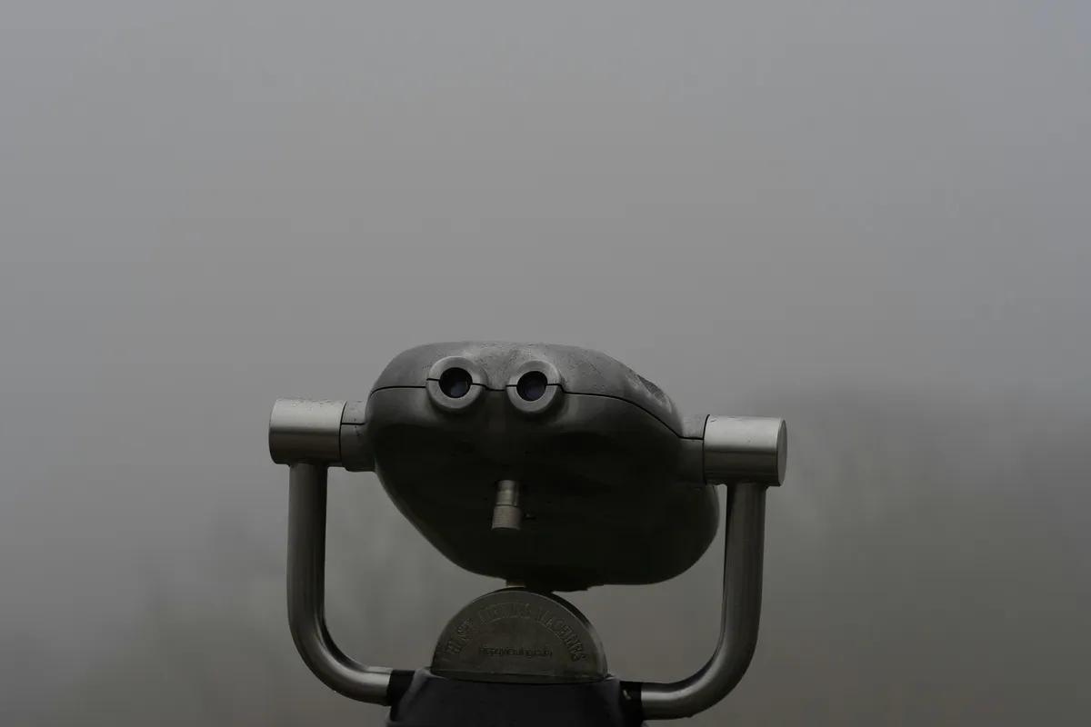 Focus Obscured by Haze A pair of gray, metal, coin-operated tower viewers on a stand. The background behind them is completely obscured by a thick, dense fog, making it impossible to see into the distance.