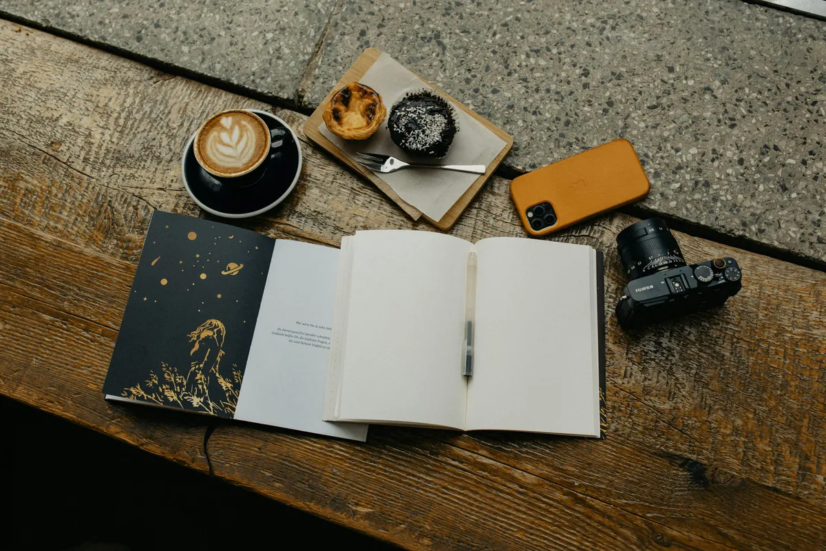 Writer's desk with grimoire and coffee A flat lay of a rustic wooden table with an open notebook and pen at its center. Surrounding the notebook are a cup of coffee with latte art, two small pastries on a board, a yellow smartphone, and a black camera. The notebook's cover has a celestial design with stars and planets.