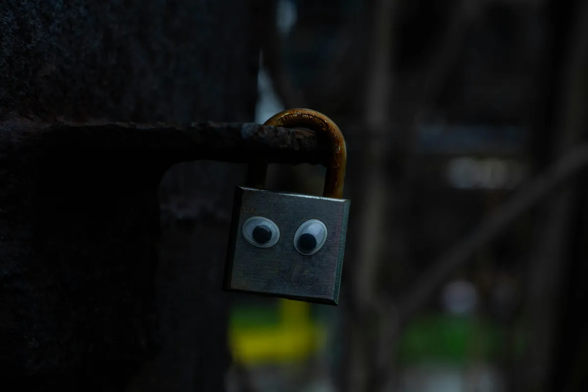 Personifying Paralysis A close-up of a square padlock with large googly eyes stuck on its front, making it look like a face as it hangs from a rusty metal hasp.