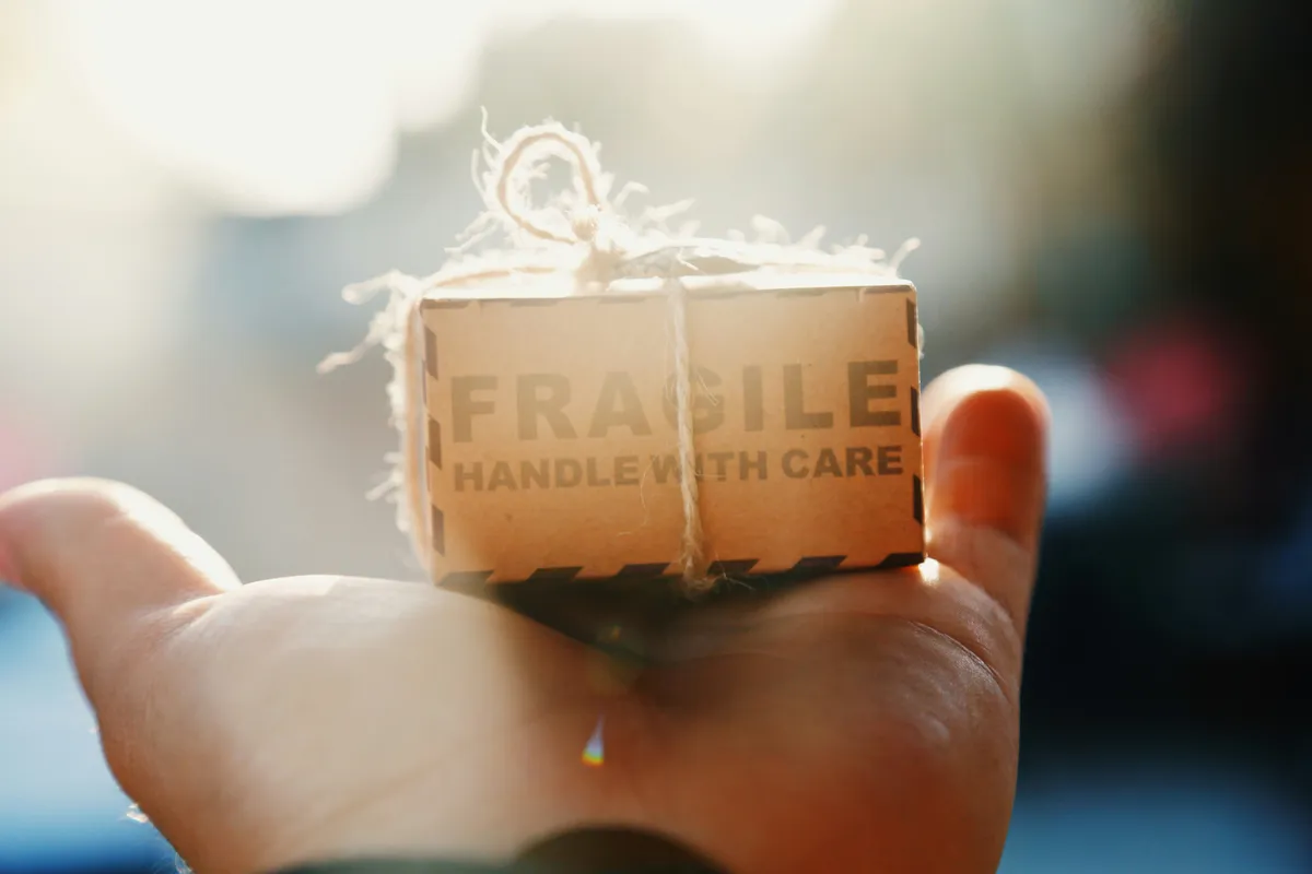 A gift for your Future Self. A person’s hand gently holds a tiny cardboard box labeled 'FRAGILE HANDLE WITH CARE,' backlit by warm sunlight, symbolizing the precious and delicate nature of our time and the importance of handling ourselves with kindness.