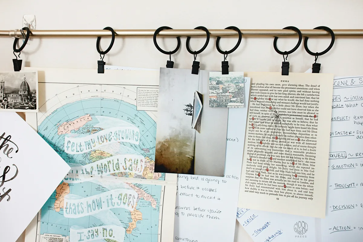 Photo A collection of papers, maps, and images hangs from black clips on a gold curtain rod, creating a writer's mood board. The papers include handwritten notes on a world map, a page of typed text with annotations, and a whiteboard with notes on story structure.