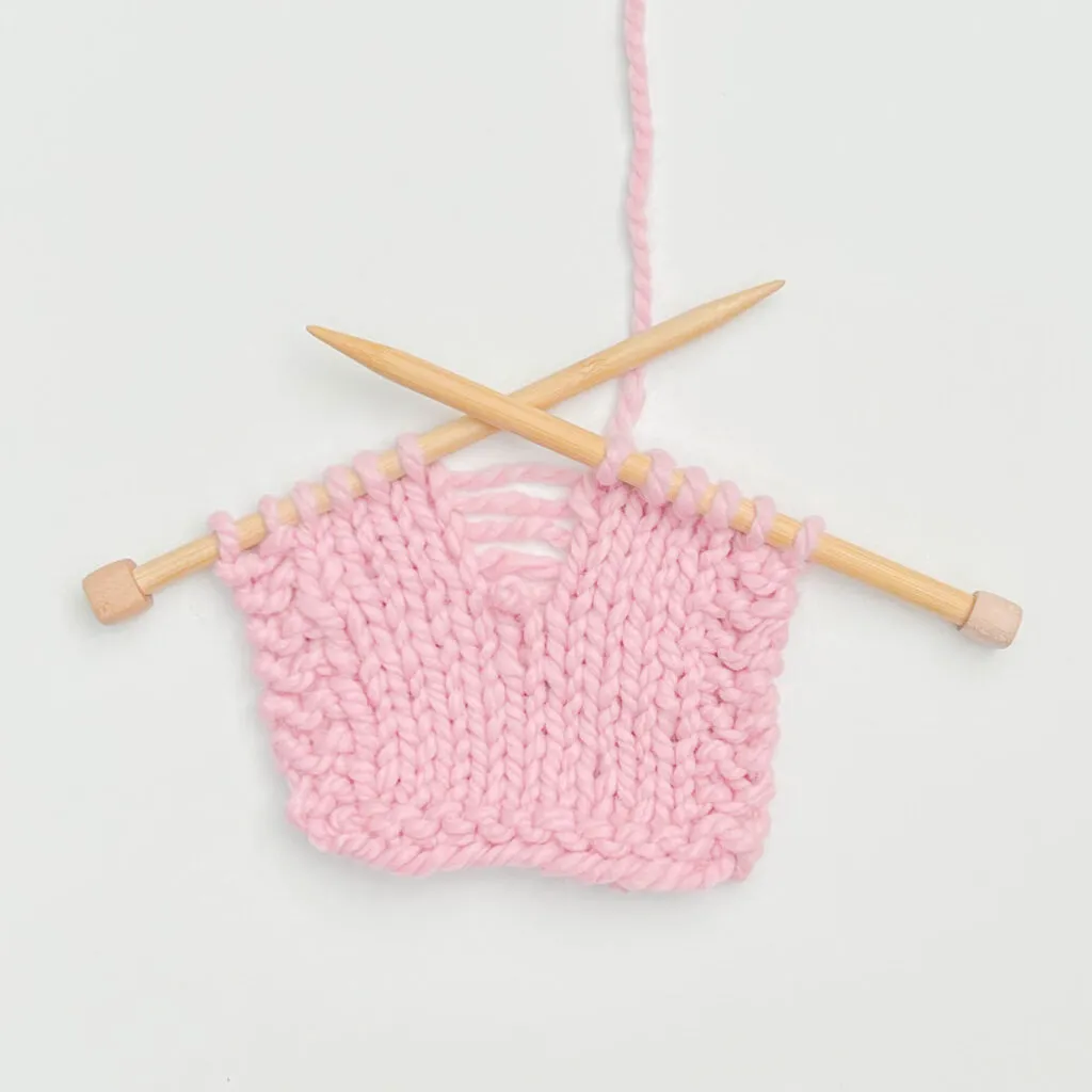A dropped stitch in a pink knitted swatch A close-up of a pink knitted swatch on two wooden knitting needles. A prominent dropped stitch in the center has created a ladder of unraveled yarn, visually representing the Stitch Drop metaphor for a working memory failure.