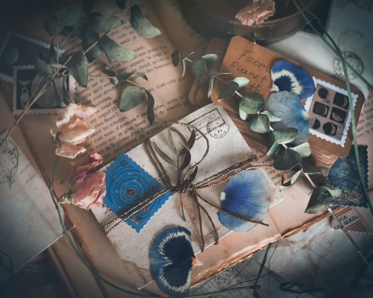 A flat lay of vintage-style correspondence against a soft, moody background. In the center, a small parcel wrapped in aged paper and tied with gold string is adorned with pressed blue flower petals and dried eucalyptus leaves. The parcel rests on a bed of old letters with cursive script, scattered with dried pink roses and decorative postage stamps. The overall mood is nostalgic and gentle.