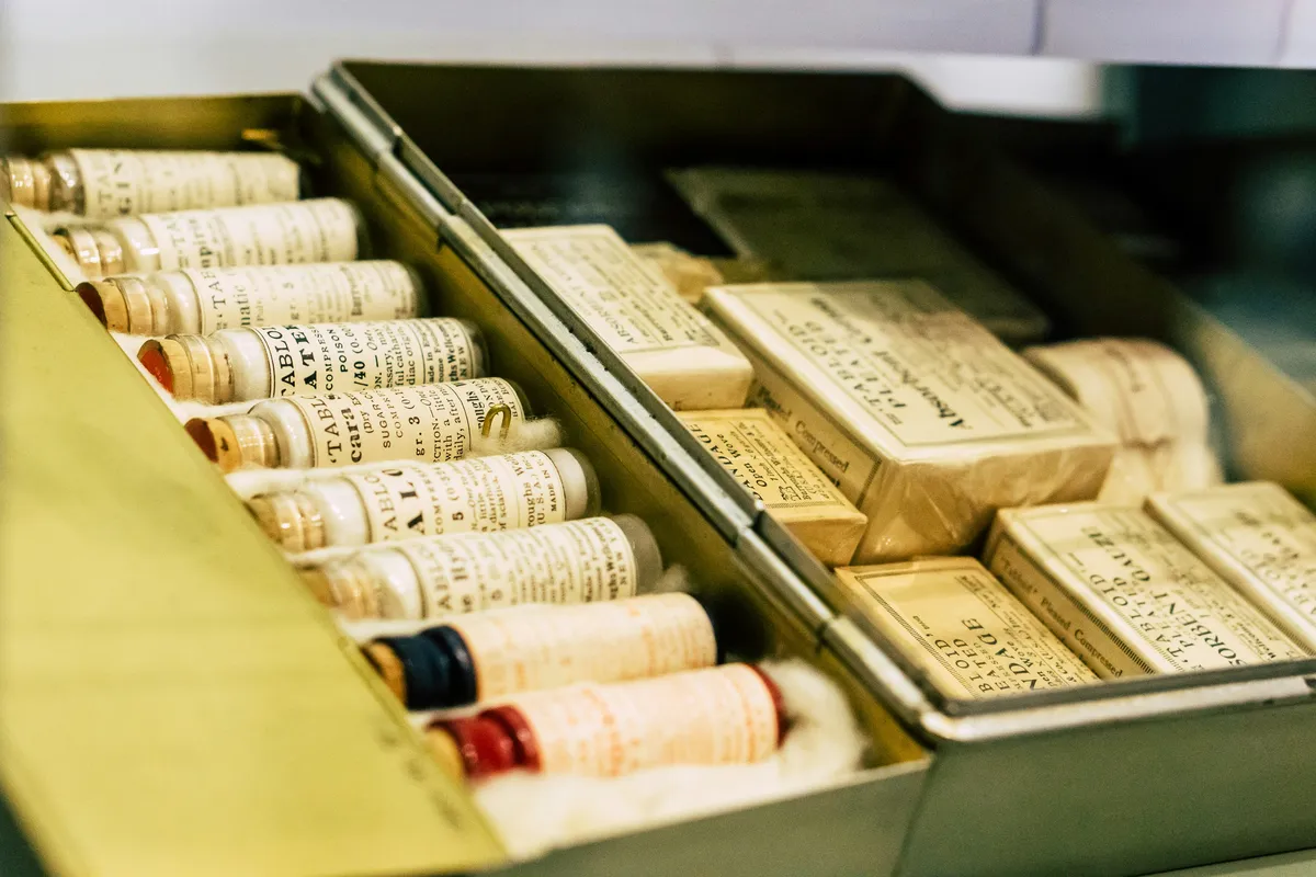 First Aid for the Mind A close-up angled view of an open vintage medical kit. The kit is lined with cotton and neatly packed with small glass vials with corks and paper labels, and old-fashioned cardboard packages of medical supplies.