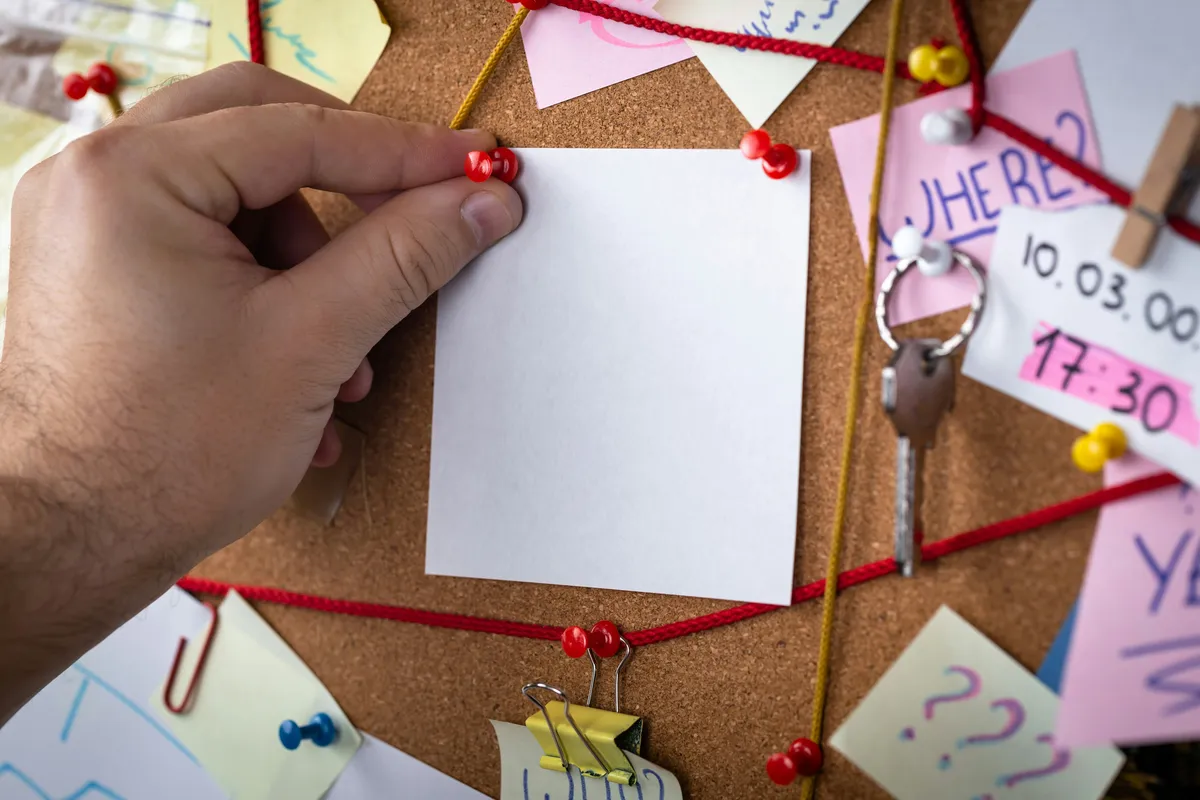 Connecting the Clues A close-up of a hand using a red thumbtack to pin a blank square note onto a cluttered corkboard that is covered with other papers and red string