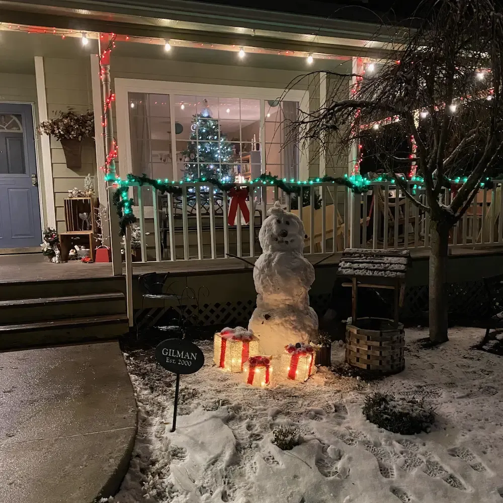 A snowman in front of our front porch surrounded by 3 light-up Christamas gifts and our Christmas tree visible through the front window.