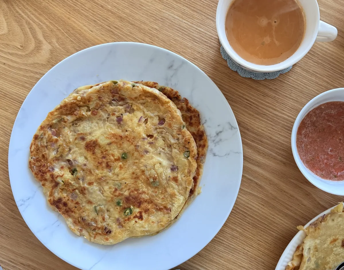 Overhead view of a weekend breakfast on a wood table: a golden pan-fried paratha flecked with red onion and green chilli on a white marble plate, a cup of milky chai on a crocheted coaster, a small bowl of red chutney, and a second plate of paratha at the edge of the frame.