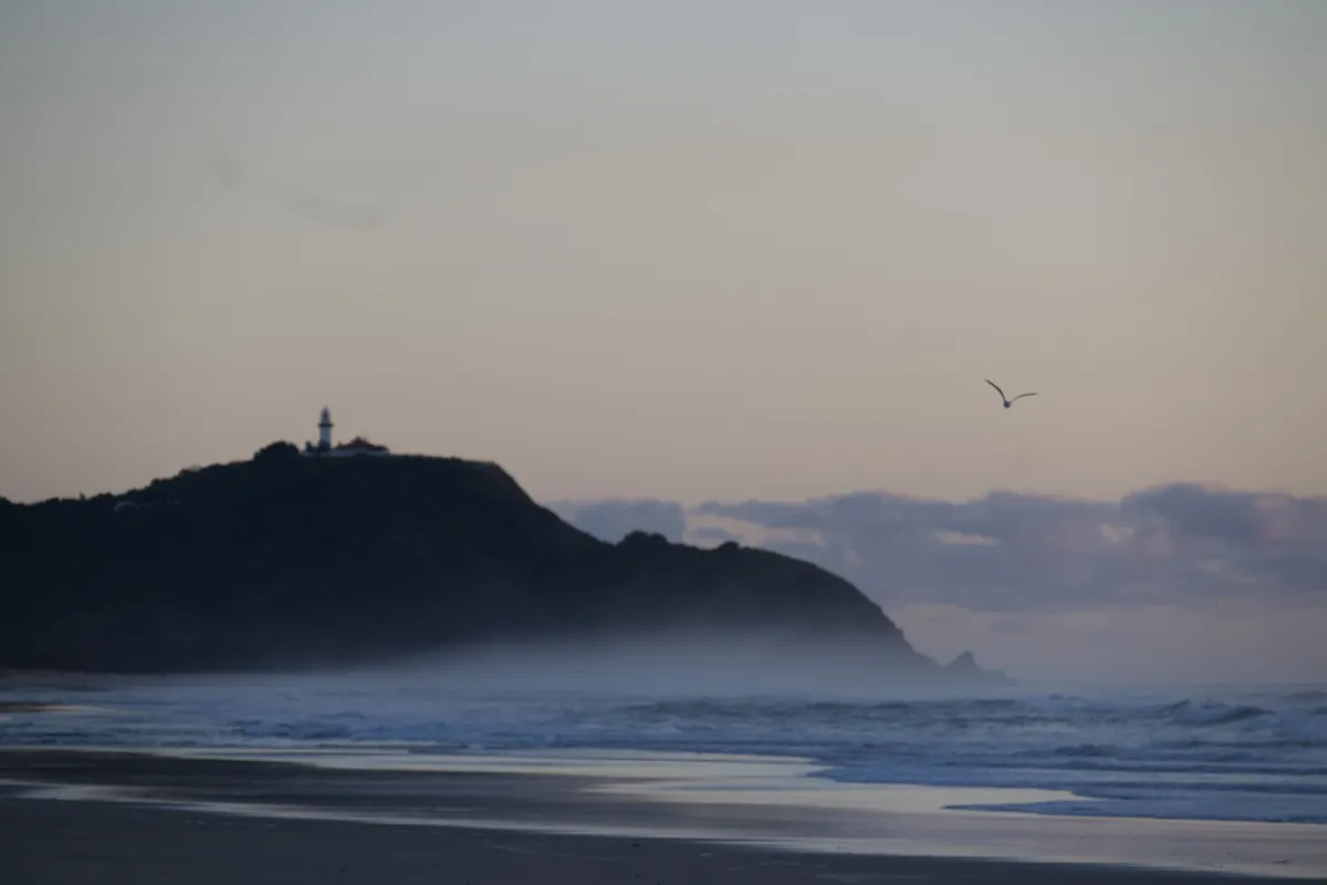 Byron Bay’s lighthouse in the distance on a dark headland above the sea, with waves crossing the foreground, low clouds on the horizon and a single bird flying through the pale sky at sunrise.