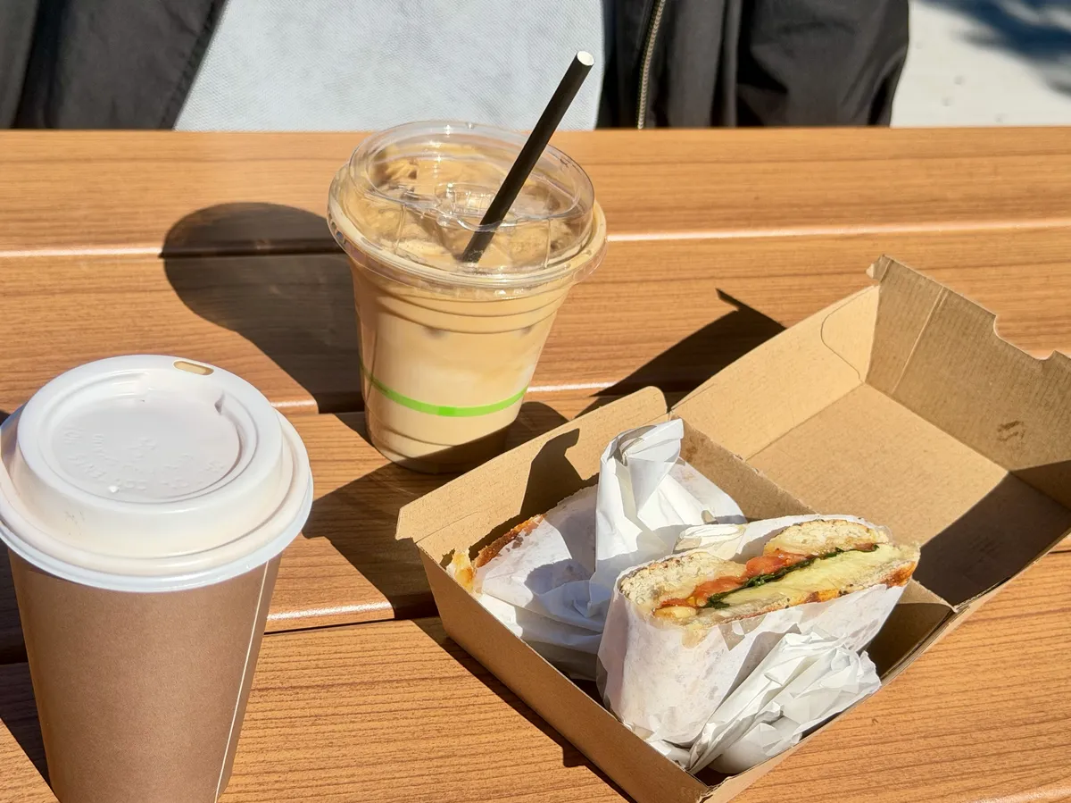 A sunlit outdoor timber table holding a takeaway breakfast: a brown paper cup with a white lid on the left, an iced coffee in a clear plastic cup with a black straw and a green band behind it, and an open cardboard box on the right with a toasted bagel cut in half and wrapped in white paper, tomato, greens and melted cheese showing at the cut.