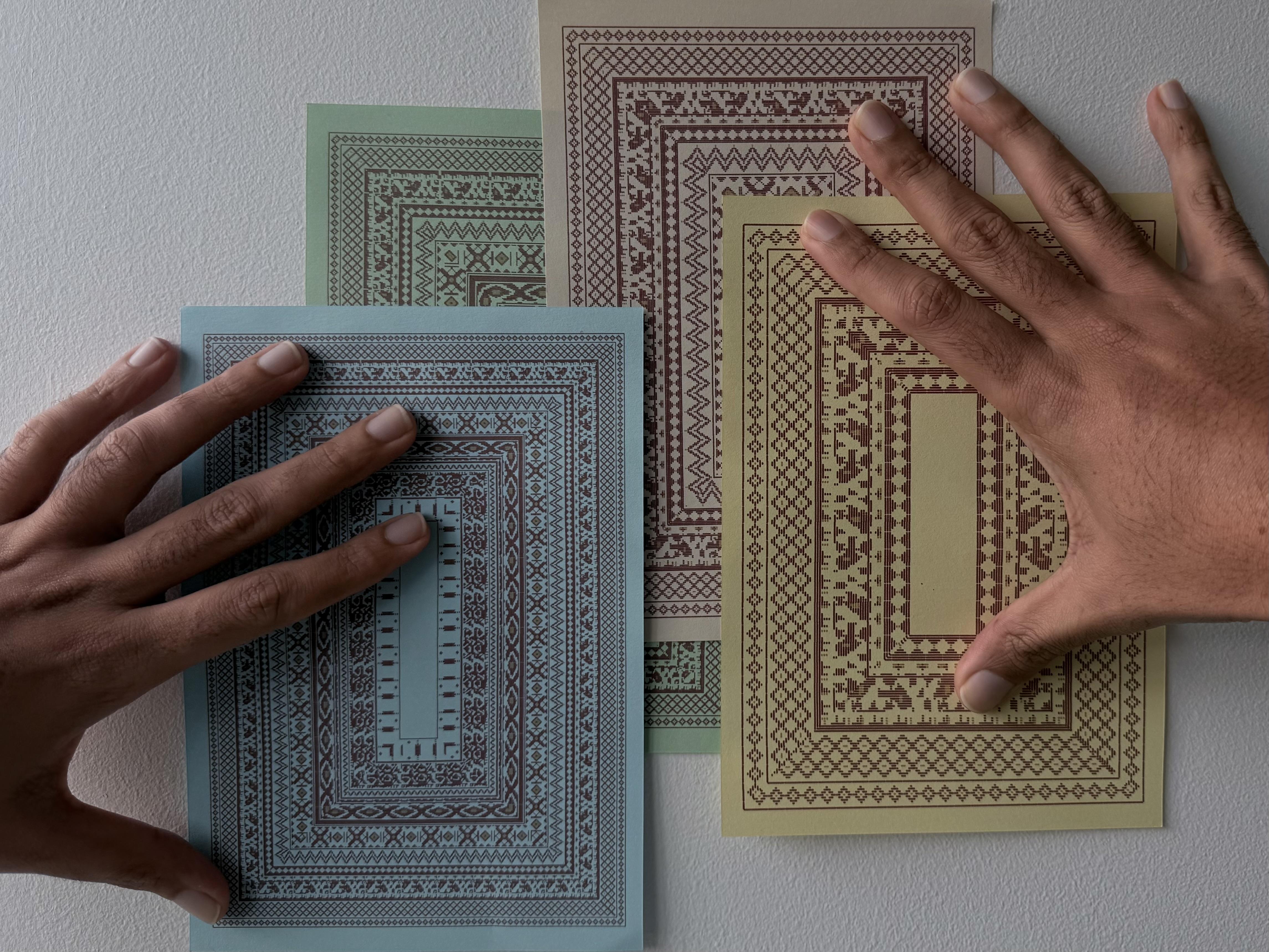 Four printed sheets of jamdani-style borders in muted blue, green, cream and grey, overlapping on a pale wall, with two hands resting on them.
