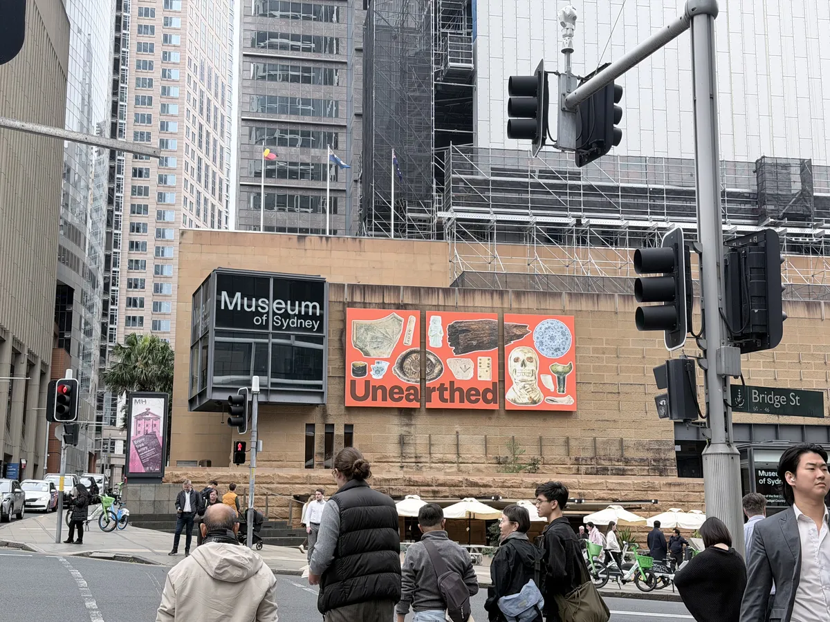 The Museum of Sydney seen from across the intersection at Bridge Street, with a large orange banner reading Unearthed mounted on the sandstone wall beside the museum entrance, office towers and scaffolding rising behind it, and pedestrians waiting at the lights in the foreground.
