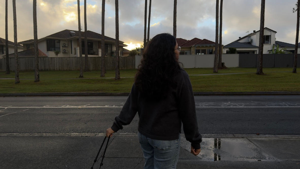 A person with long dark hair seen from behind, holding a purse handle, standing at the kerb of a quiet suburban street at dusk, facing houses and tall palms with the sun low behind cloud and a puddle on the path.