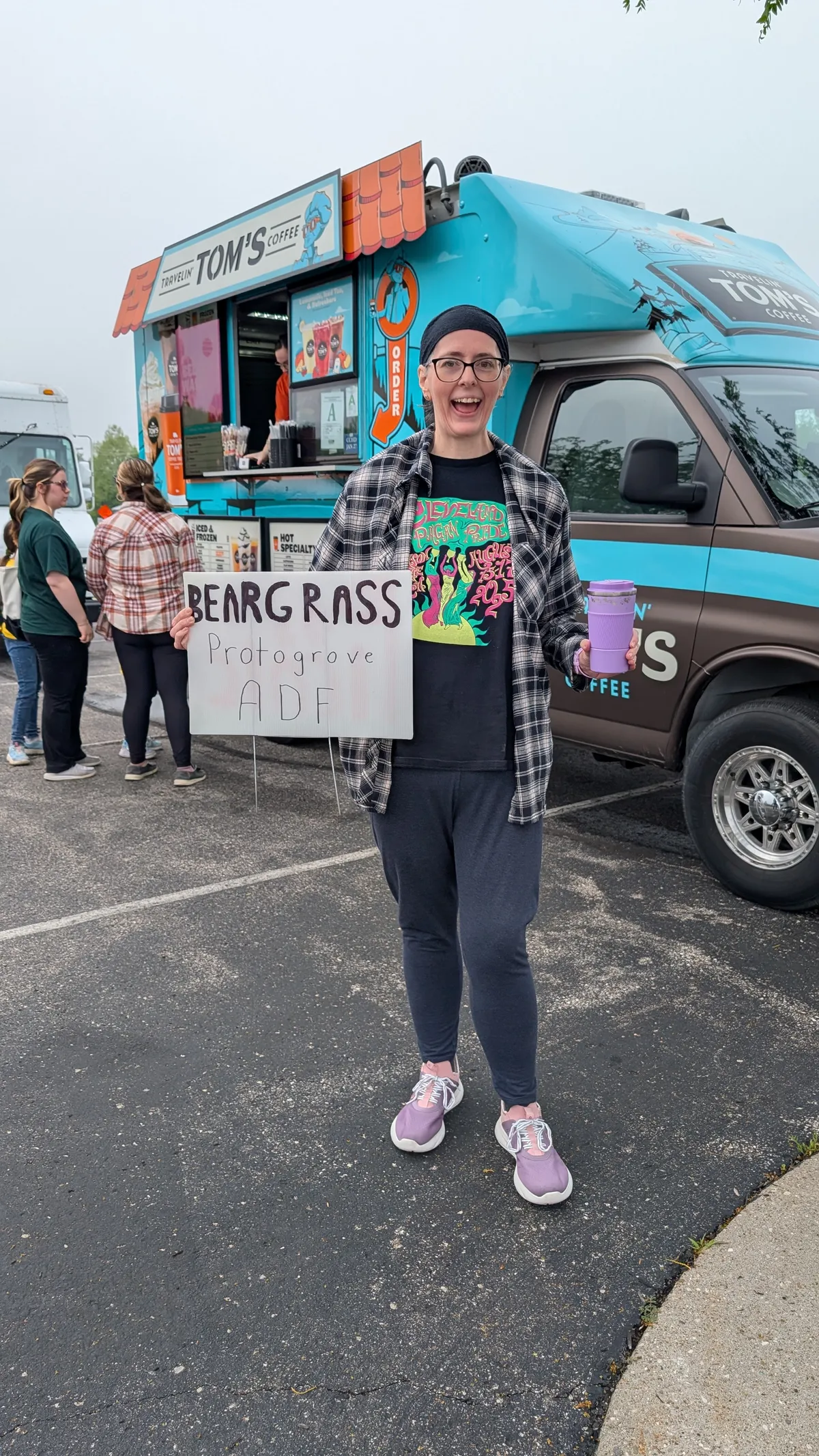 A smiling woman stands in front of a food truck, holding a cup of coffee and a sign that says Beargrass Protogrove, ADF.