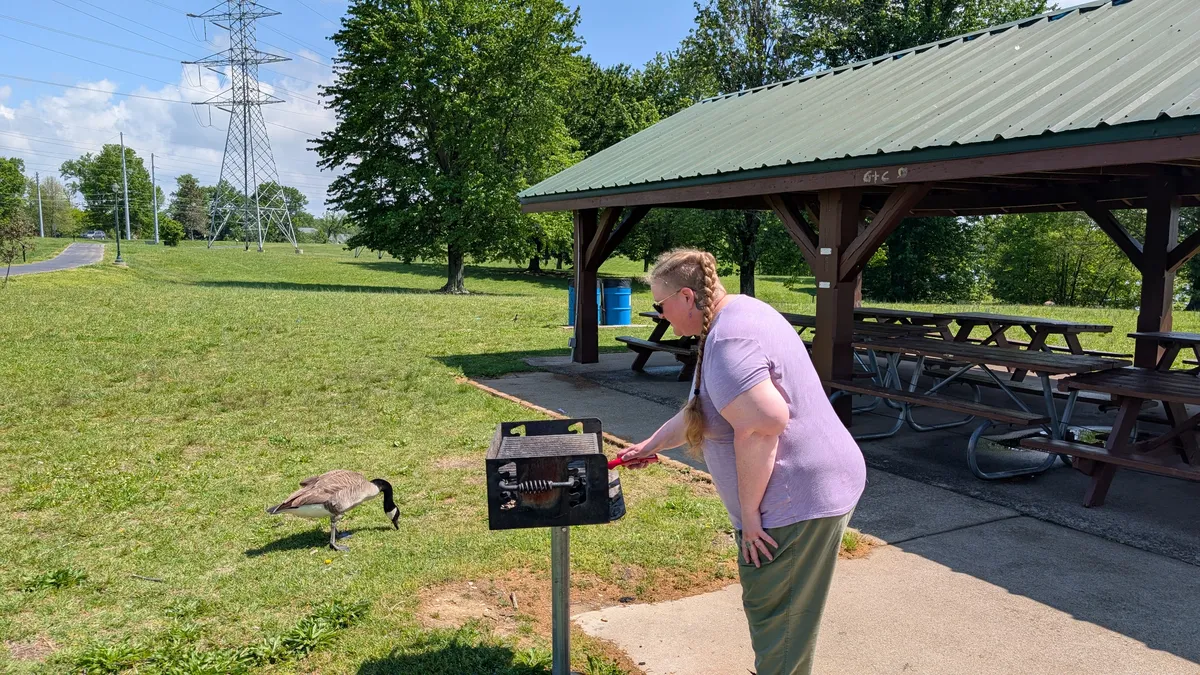 A Druid wearing a purple shirt is lighting a charcoal grill, while a Canada Goose stands nearby.