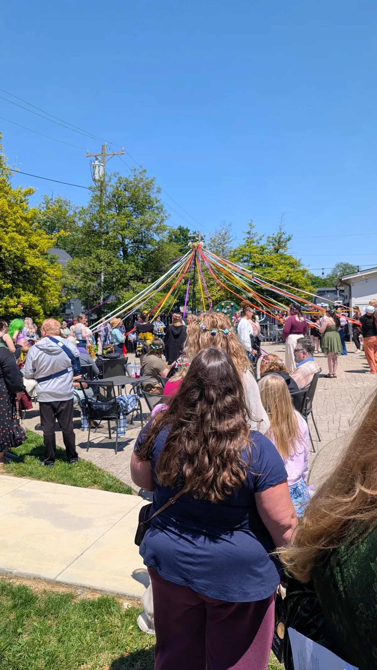 Under a bright blue sky, about 30 people stand around a Maypole decorated with a rainbow of colored ribbons. Some are holding the ribbons, preparing to weave them around the pole in a traditional Maypole Dance.