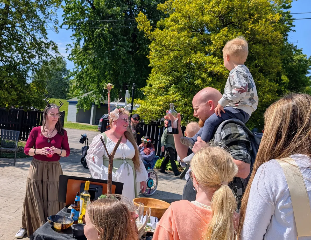 A small group of people is lined up around an altar decorated for Beltane. Two Druid people wearing dresses and antlers stand behind the altar. In front of the altar, a man with a young child on his shoulders raises a bottle, preparing to make an offering.