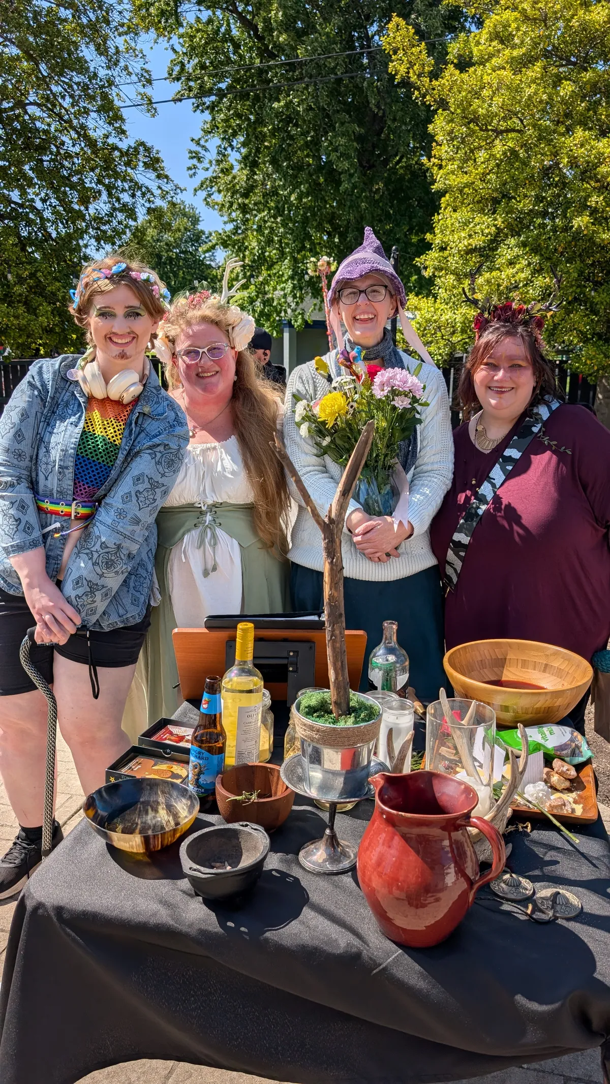 Four smiling Druids stand behind the altar. The altar is decorated for ritual with a branch standing in a pot and several jars, bowls and pitchers.