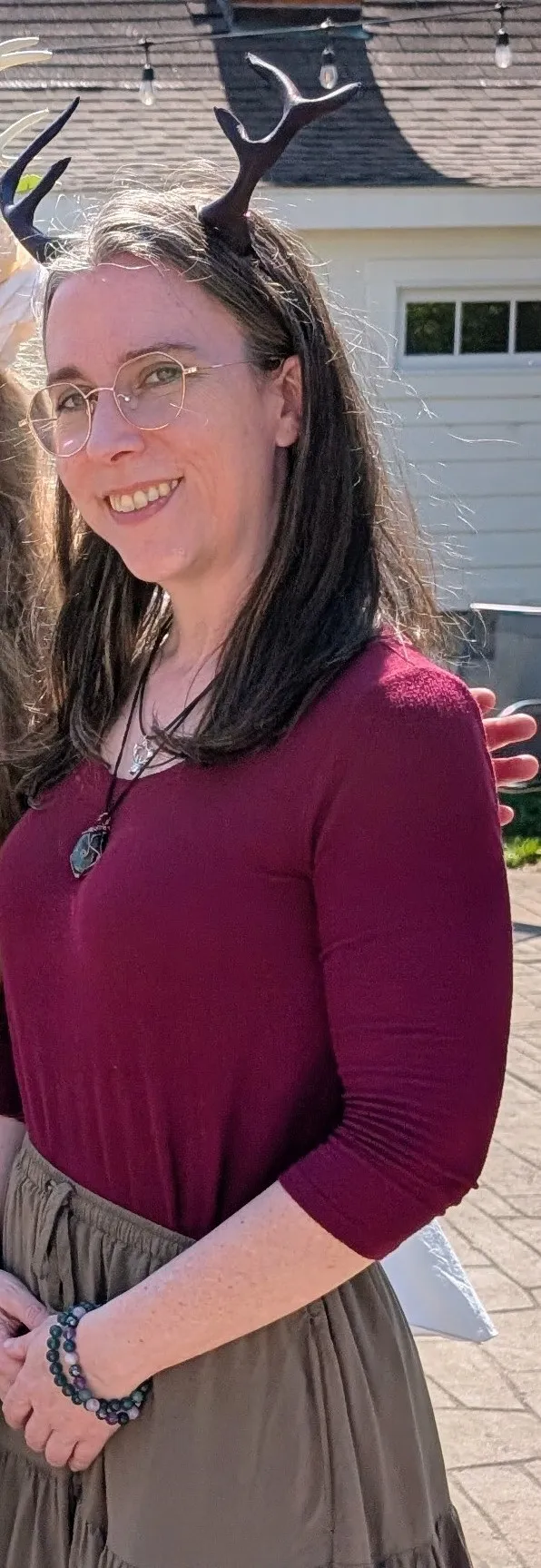 A smiling Druid with long brown hair, wearing antlers, a gemstone necklace and a maroon shirt.