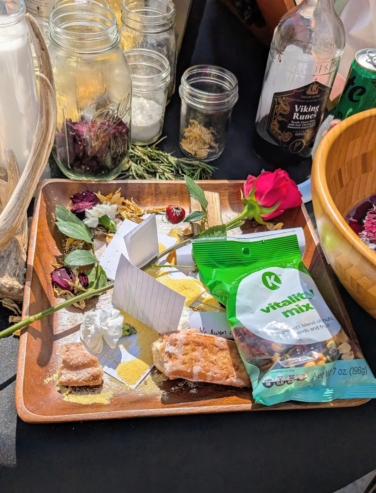 A bamboo plate covered in offerings: a bag of trail mix, two carnations, some rose petals, cornmeal, a snack cake, a small piece of red heart-shaped jewelry.