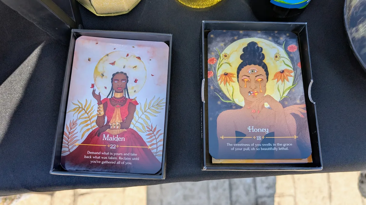 Two cards: The Maiden, which depicts a dark skinned woman wearing gold jewelry and a red dress with the Full Moon behind her; and Honey, which depicts the face of a dark skinned woman whose eyes drip with golden tears of honey.