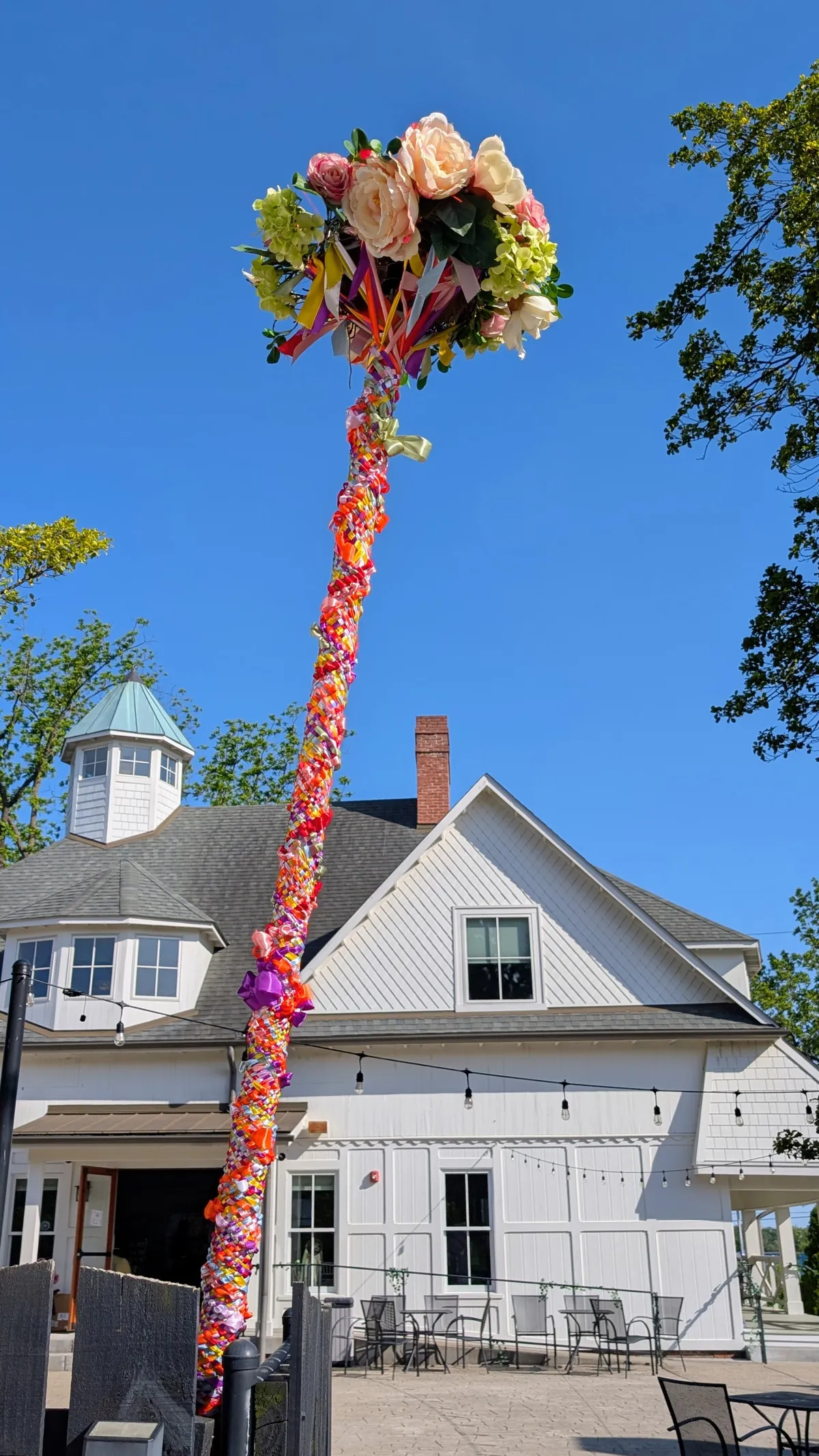 A Maypole stands in front of the Carriage House, under a bright blue sky, covered in colorful ribbons that were woven around it during the Maypole Dance. There is a lovely bouquet of flowers at the top of the pole.