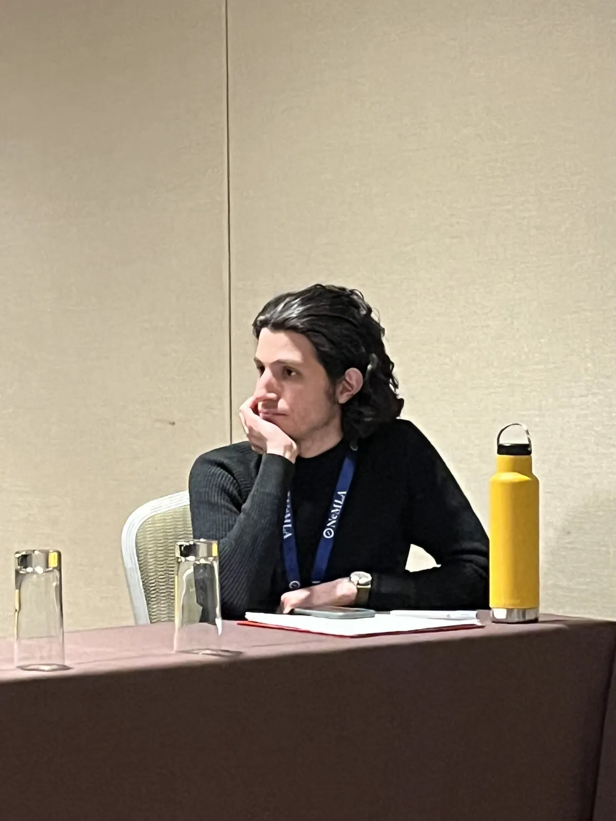 A man in a black sweater sits at a conference table gazing to his right