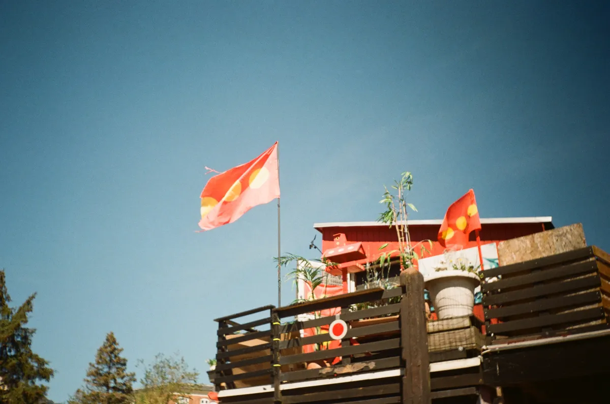 A picture of a red building with a balcony, containing pot plants. The balcony is flying a red flag with three yellow circles arranged horizontally across it. The building is photographed against a blue sky.