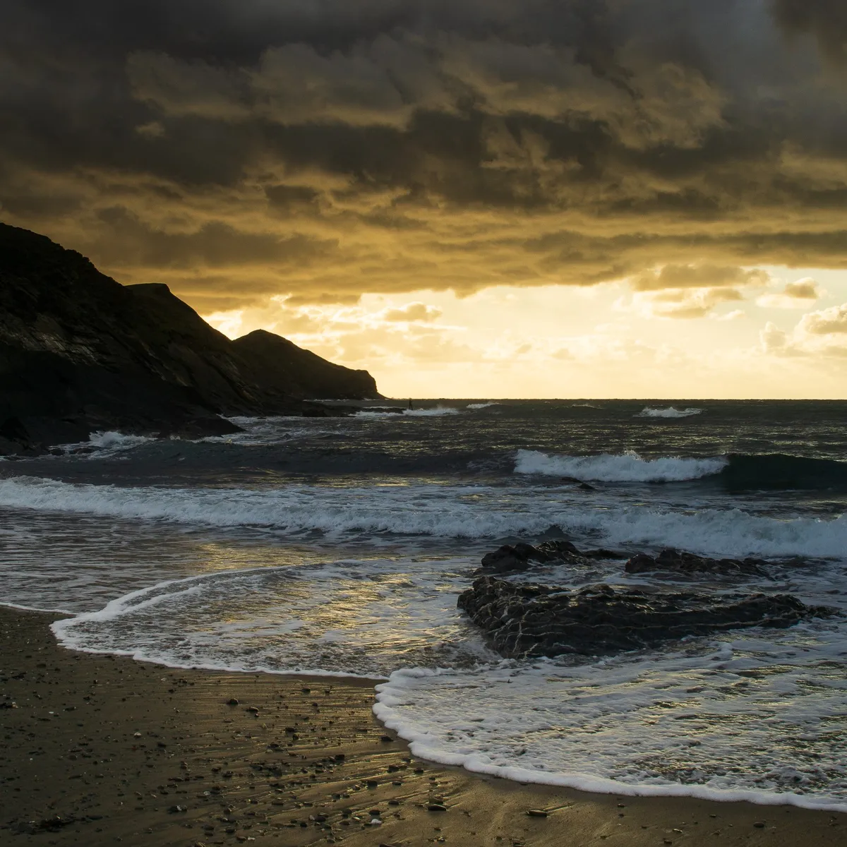 Evening beach scene looking out over the sea, a golden slither of evening light illuminates the incoming tide forming shallow aprons on the sand closest to us. The headland and rocks are silhouetted on the left (a sea angler is just visible, casting from a scary-looking distant spot on the rocks) and a mass of gathering uplit clouds above the golden area of sky look forebodingly dark.