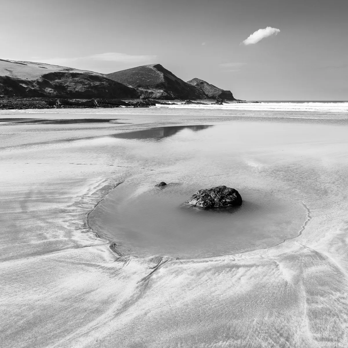 In black and white, a sandy beach stretches away from us wide and open to the sea, with rocky headlands on the left. A single rock sits in a depression in the sand, surrounded by a moat of sea water. The sky above is clear except for one little fluffy cloud