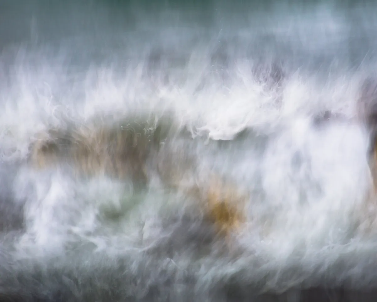 long exposure and camera movement give a very blurred and energy-filled view of a wave crashing over a rock, with streaks of white, blues and sea greens
