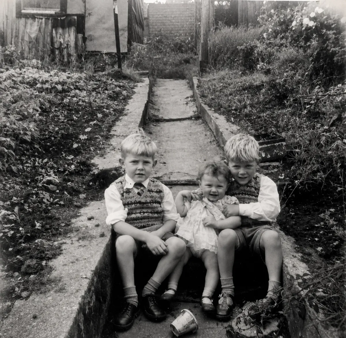 Two 5 and 6 year old brothers and their 2 or 3 year old sister sitting on the back step dressed in their Sunday best in the late 1950s.