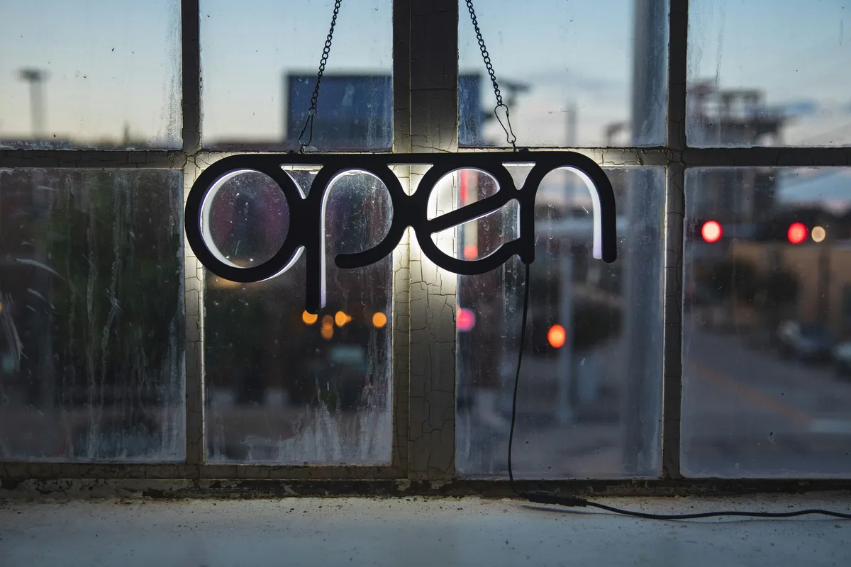 An open sign hanging in a window with a blurry city behind it.