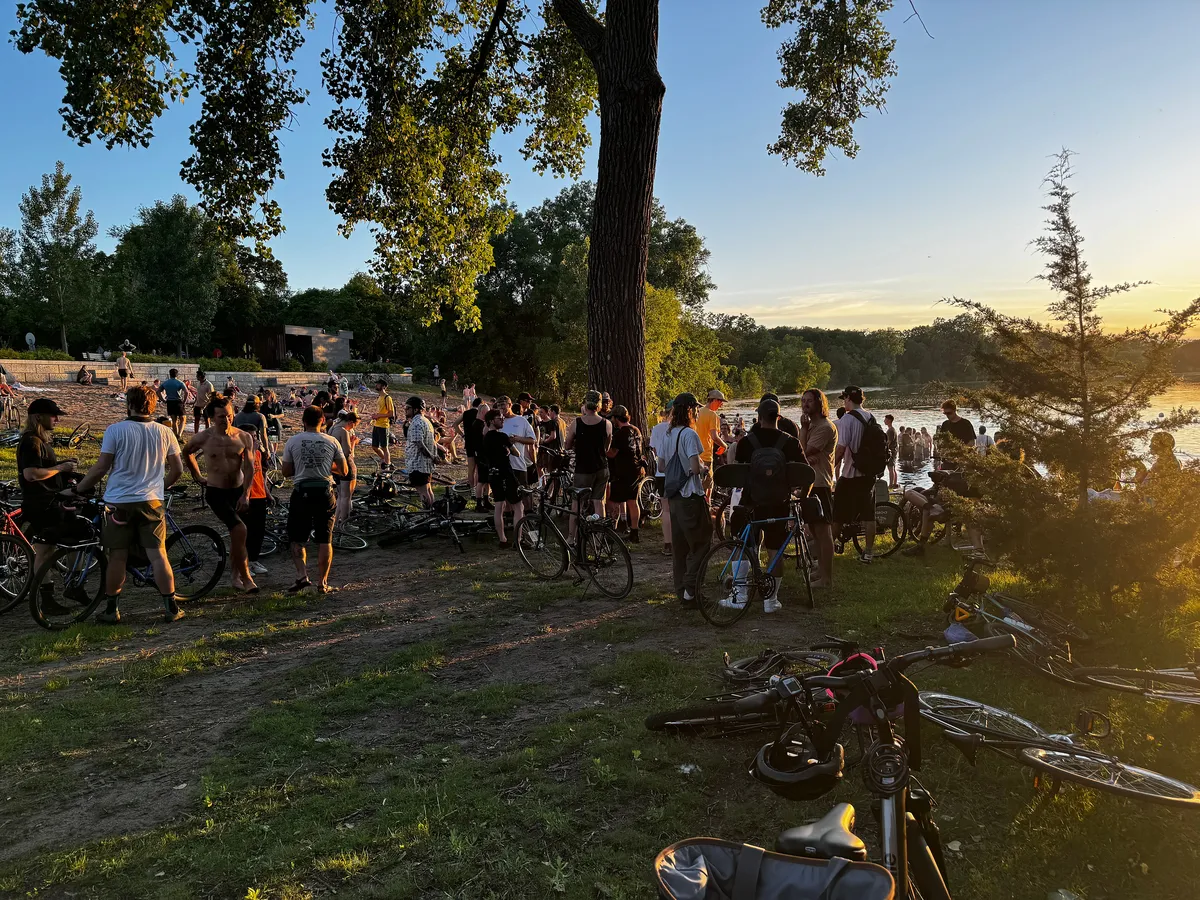The sun setting on a beach with people standing around talking with bicycles laying in the foreground.