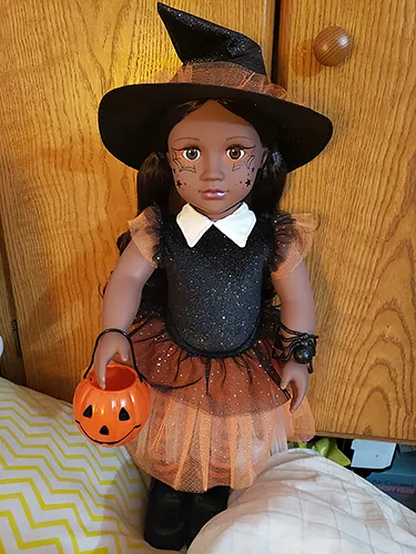 A dark-skinned, dark-haired doll dressed in an orange an black, sparkly witch costume, complete with spider bracelet. She's also holding a Trick-or-Treat jack-o-lantern bucket.