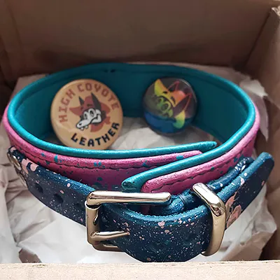 a pink and teal leather collar with reverse color paint splatters and shiny nickel hardware. In the background are two pinback buttons featuring High Coyote Leather's mascot, one in brown and black and the other in a rainbow gradient