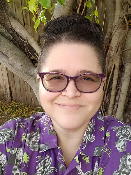 a selfie of the creator: a light-skinned individual with a dark brown fauxhawk, purple square frame glasses, and wearing a Beetlejuice print button-up shirt. Behind them is a tree covered in vine-like roots.'