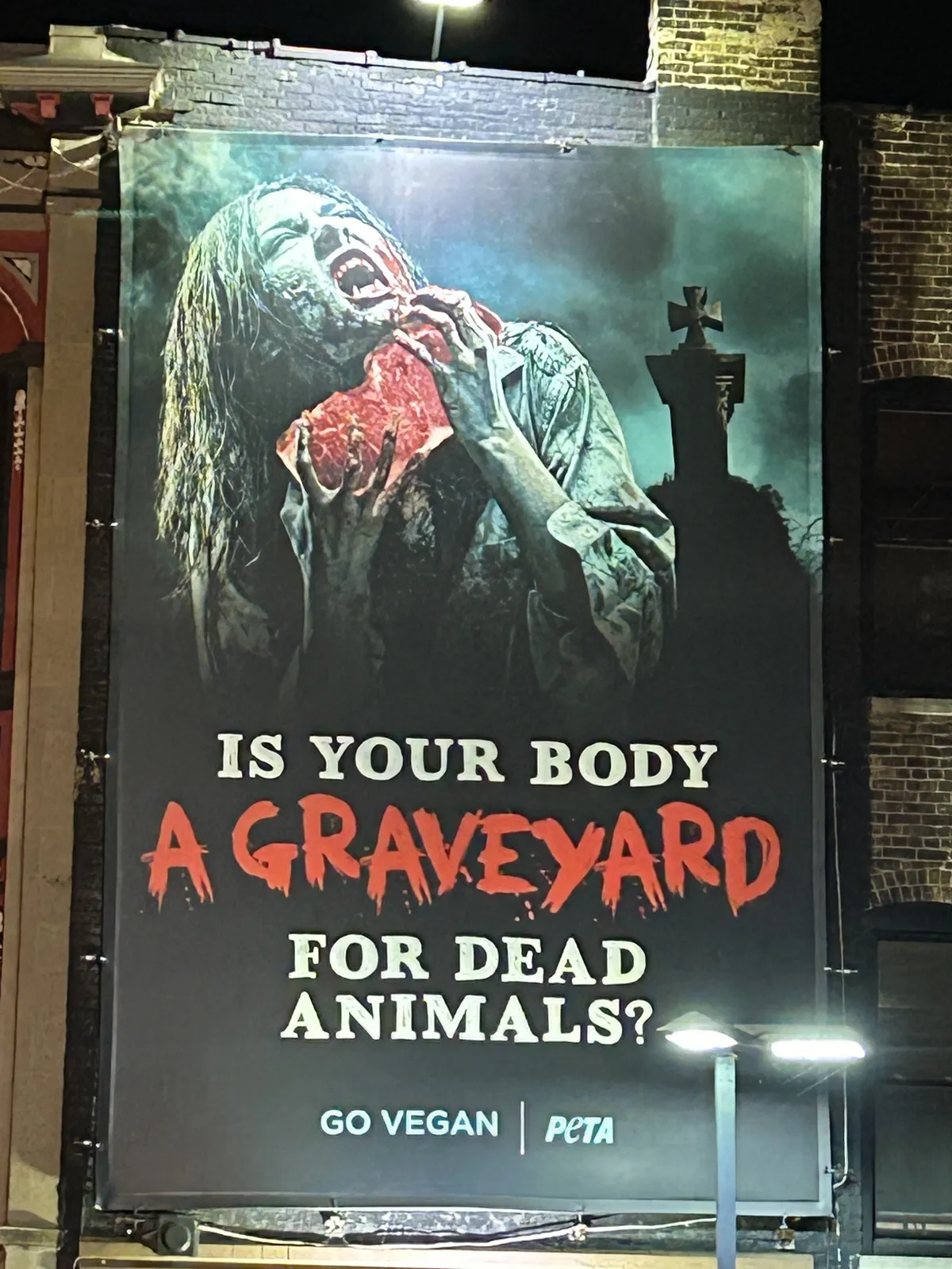 A billboard illuminated at night showing a person eating bloody meat above the words "Is your body a graveyard for dead animals?"