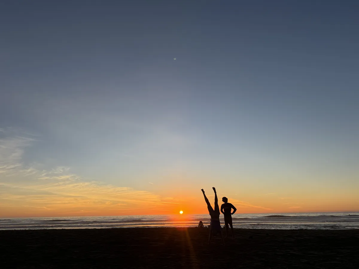A person attempting a roundoff on Ocean Beach at sunset while a friend watches.