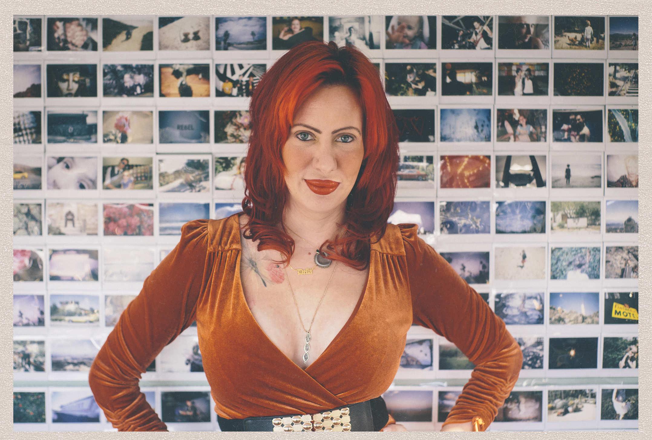 A ridiculously attractive woman with red hair and an orange v-neck velvet dress posing in front of a wall of instant film photos