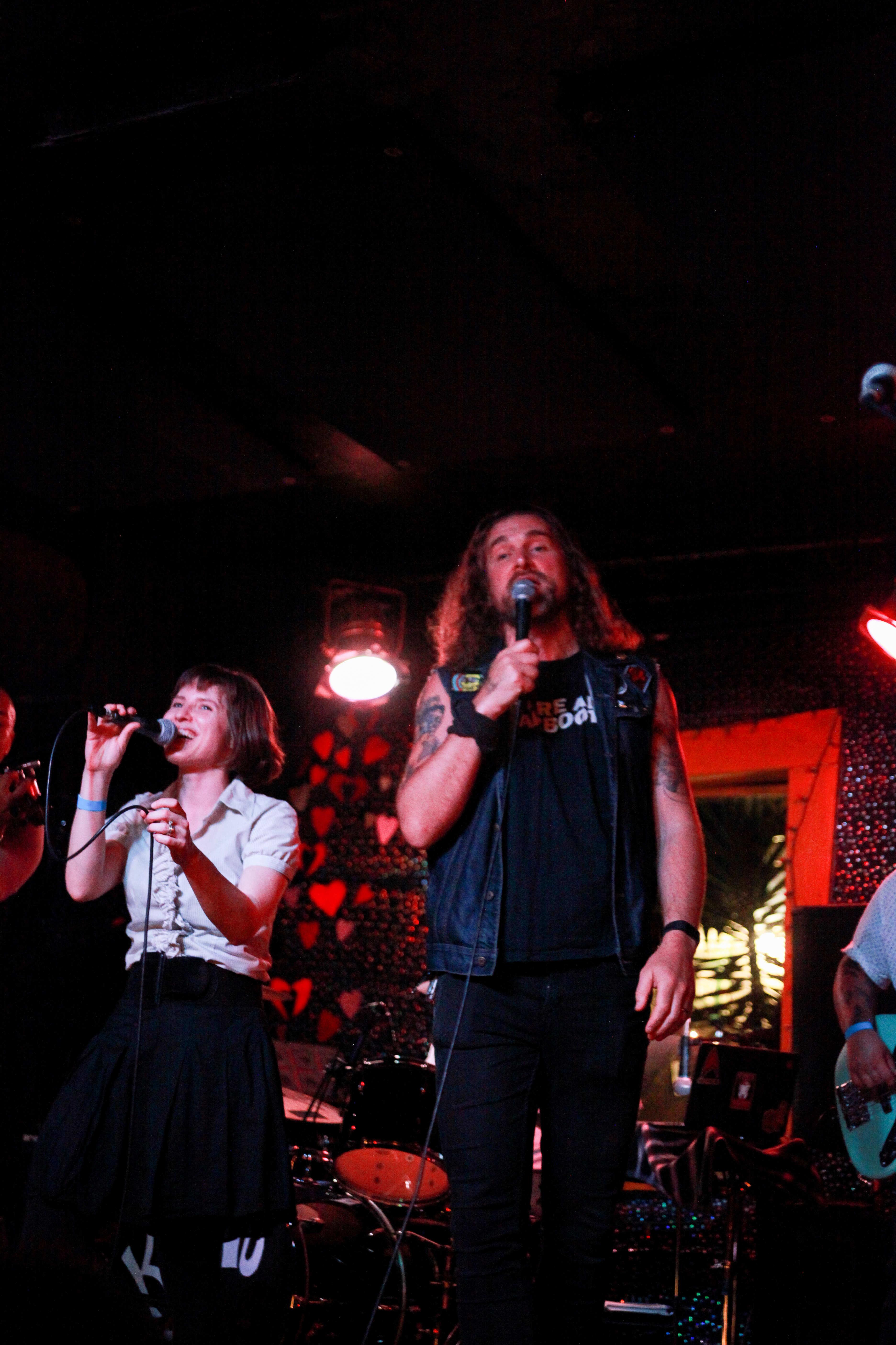 Two people singing on stage; a tall man with glorious jesus hair and a denim vest, and a woman in a greyish blouse and black skirt