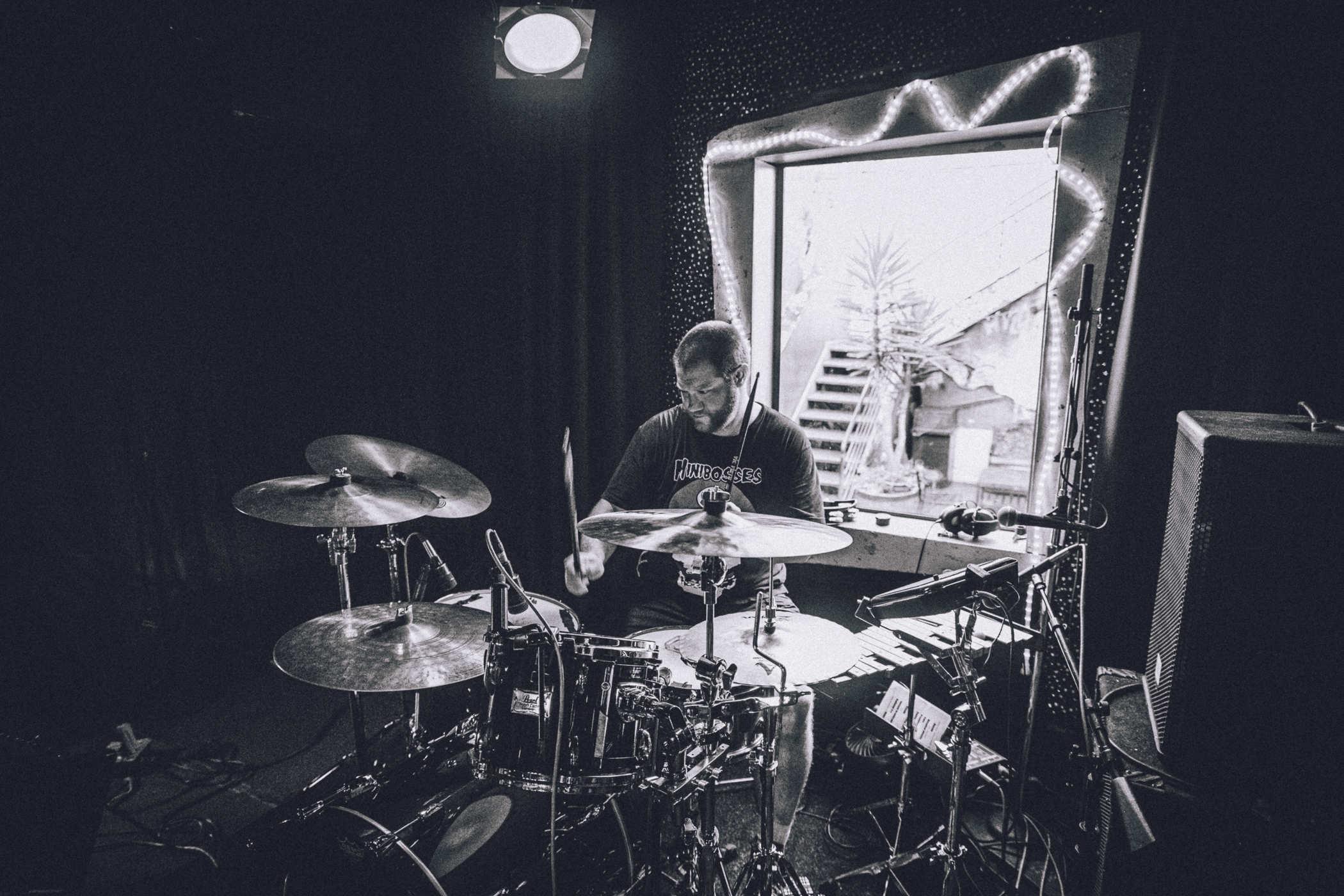 Black and white photo of a person setting up drum equipment on a stage in front of a window with ornate tube lights around it