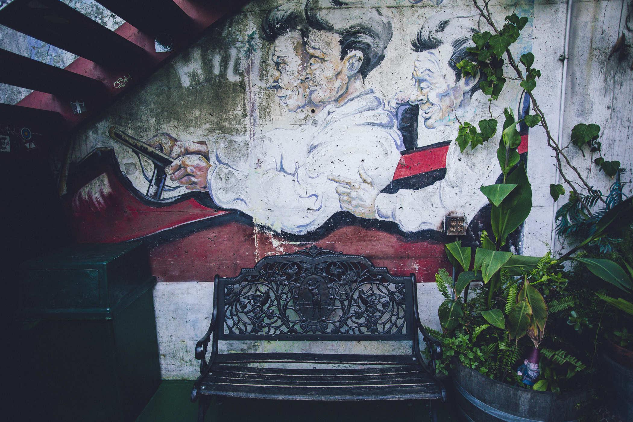 Photo of an outdoor patio with a bench under stairs; a mural on the wall depicts three people in a car