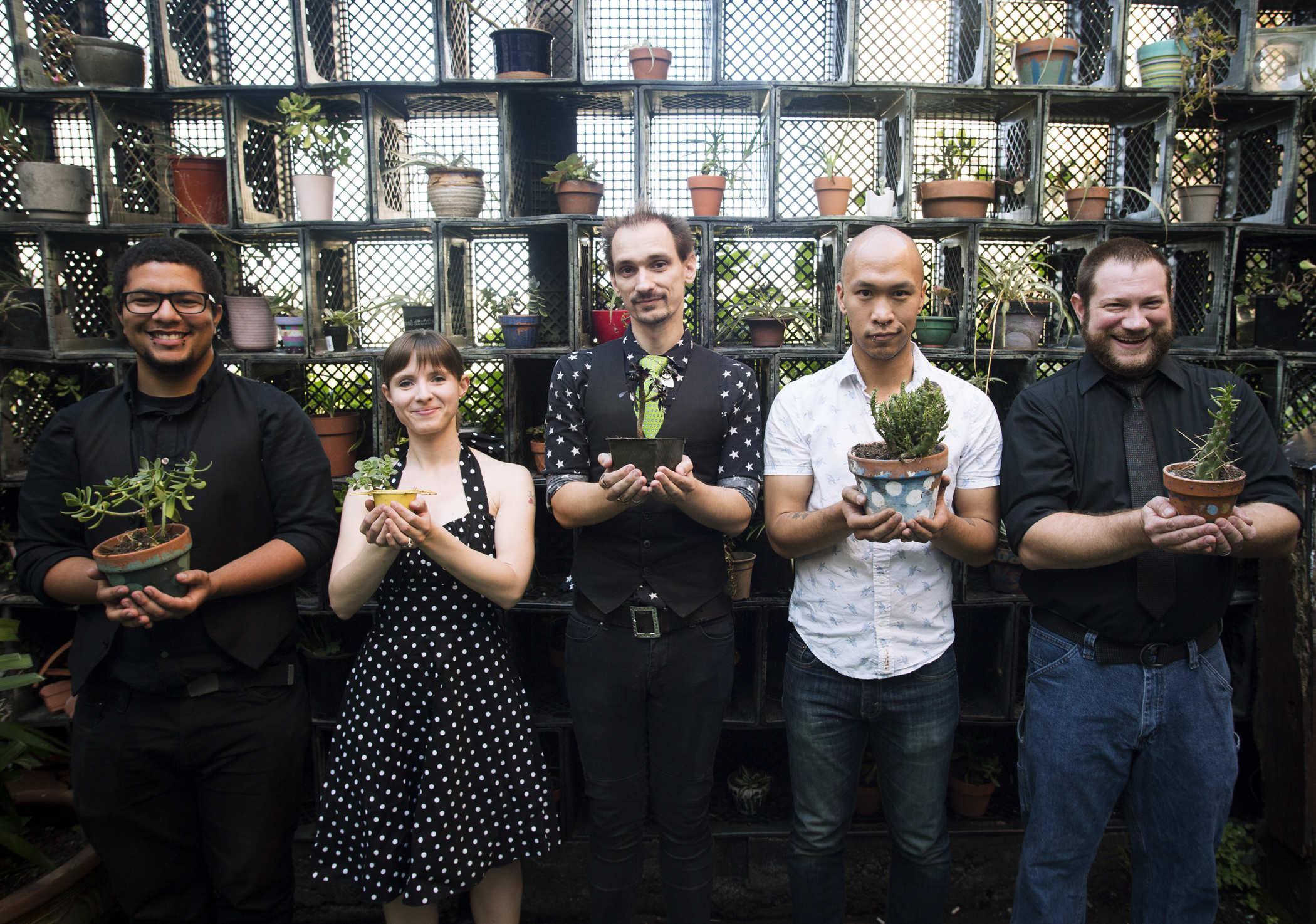 Photo of five people standing in front of a wall of milk crates containing succulents; each person is holding a small potted plant and looking at the camera