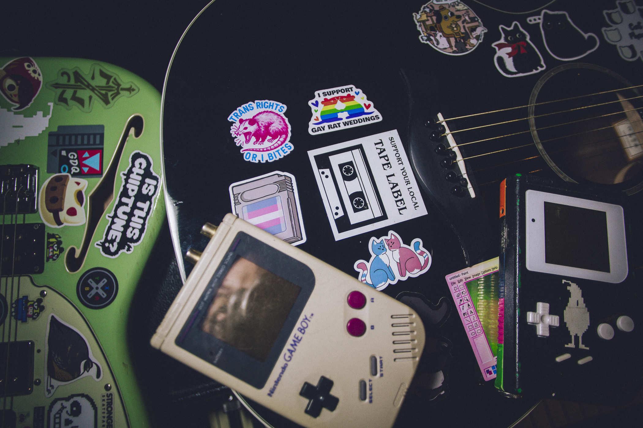 Photo of two nintendo gameboys (one painted black with a white quail image on it) on top of a black, sticker-covered acoustic guitar many of which call for trans  rights; there's a greenish electric guitar next to it with a hard to miss 'IS THIS CHIPTUNE' sticker
