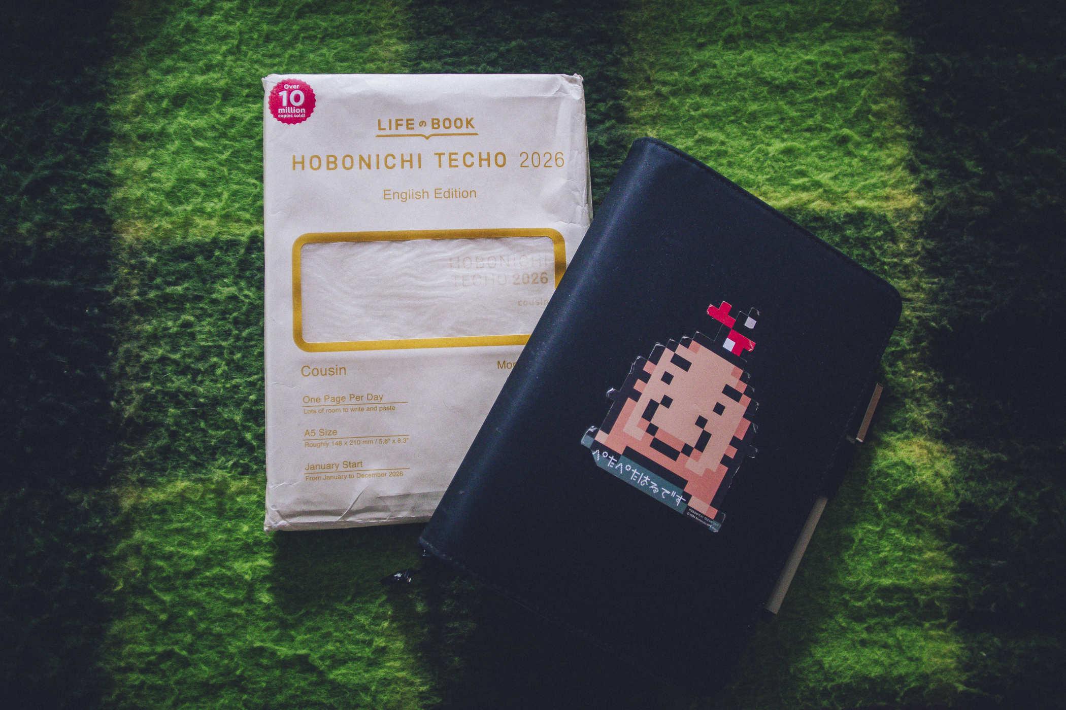 Photo of a hobonichi techo cousin english 2026 planner, still in its packaging, next to a large black cover with a Mr. Saturn character from the Mother video game series on it