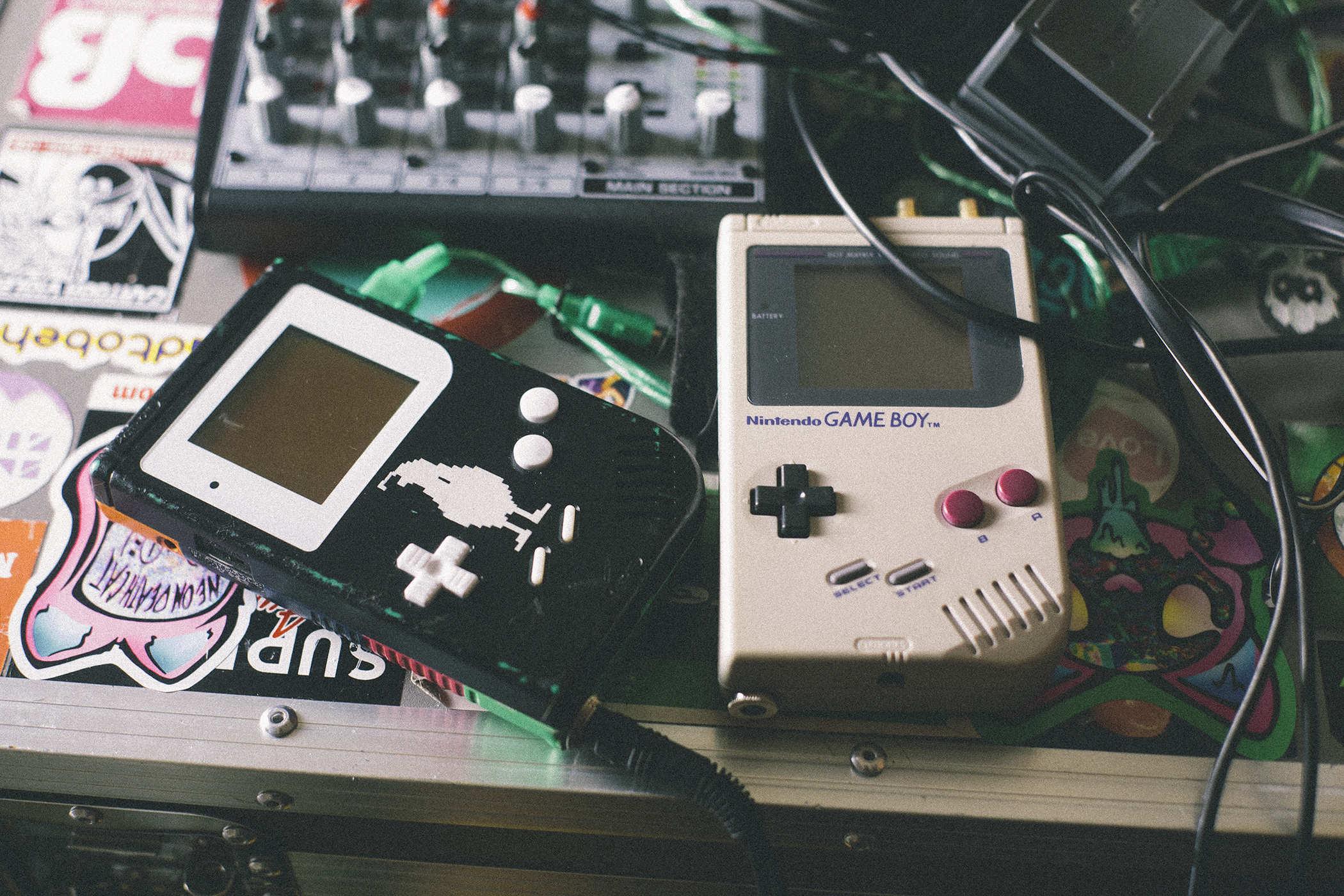 Photo of two nintento Gameboys rigged up to play music with on top of a container covered in stickers