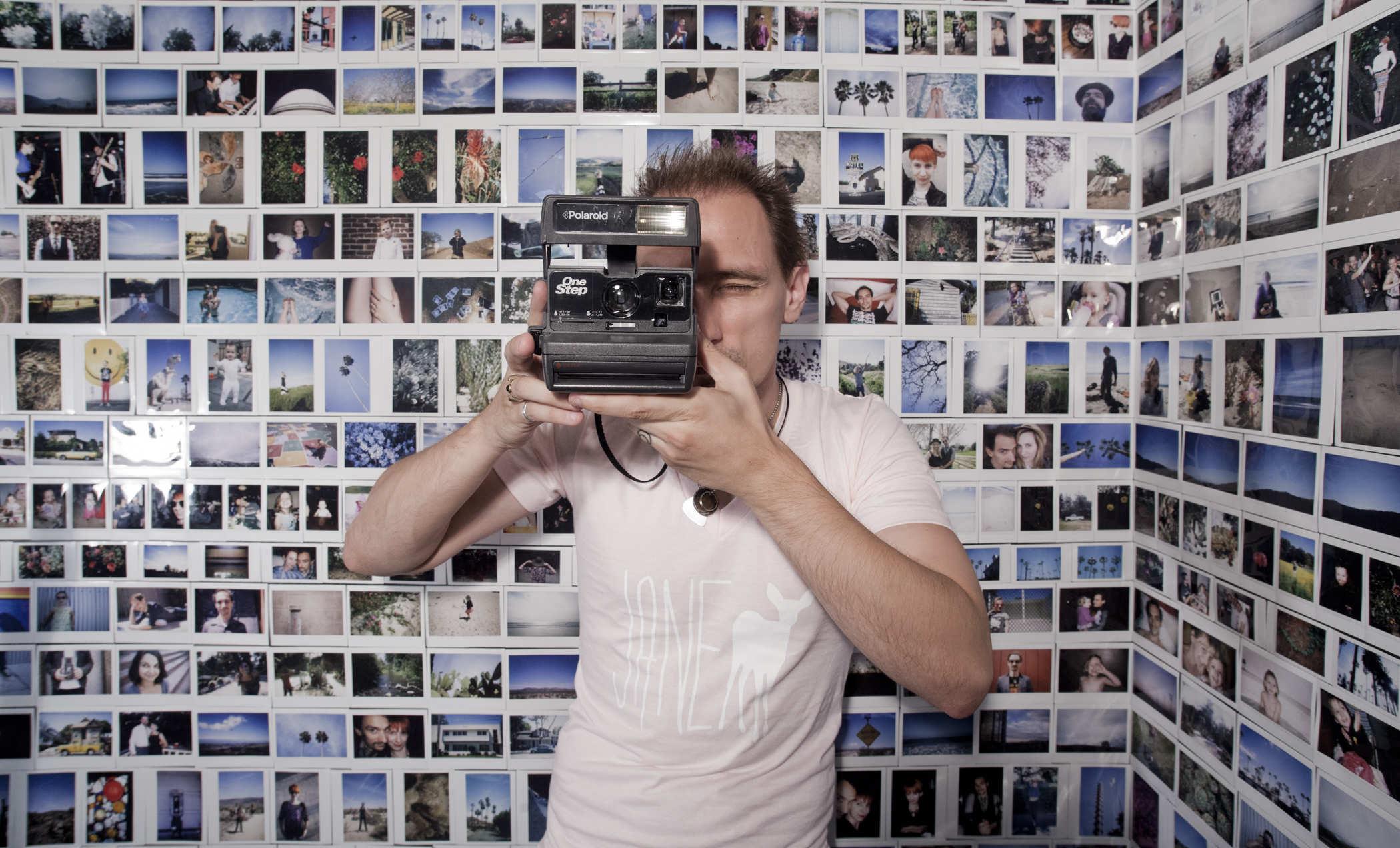 Photo of a person wearing a pink shirt with a white deer and the word 'JANE' holding up a polaroid camera; they're standing in front of a wall COVERED in instant film photos