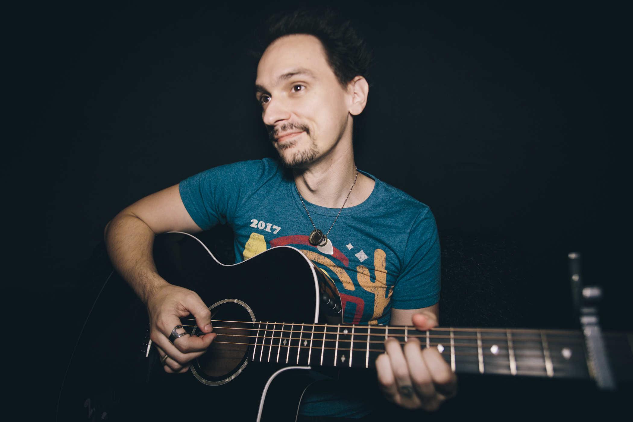 Me wearing a blue-green 'Desert Bus' shirt holding a black acoustic guitar on a black background
