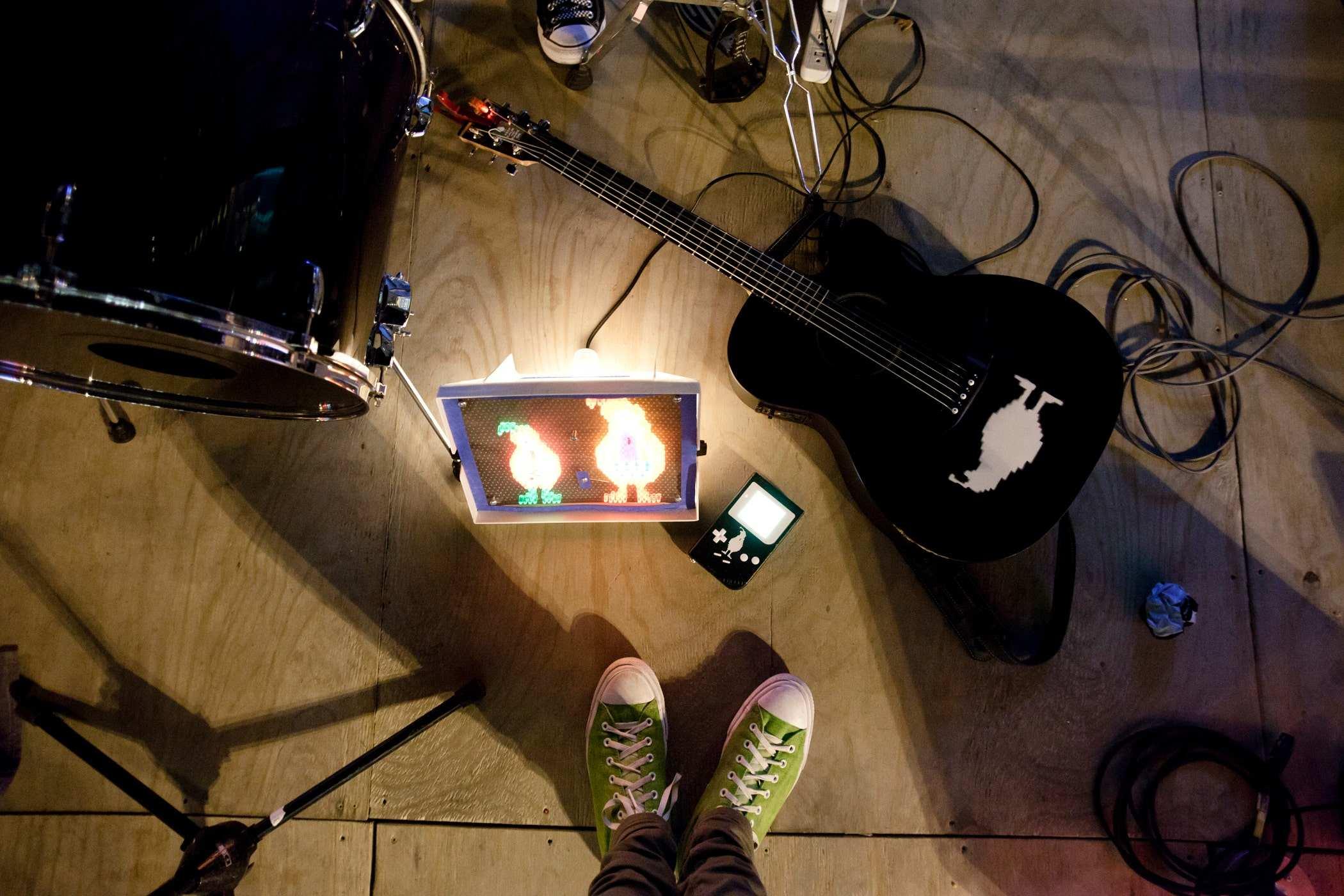 Photo of my shoes taken on a stage; a black acoustic guitar with an image of a quail is on it, as well as a nintendo gameboy, a kick drum, some cables and a lite brite toy with the same quail logo on it