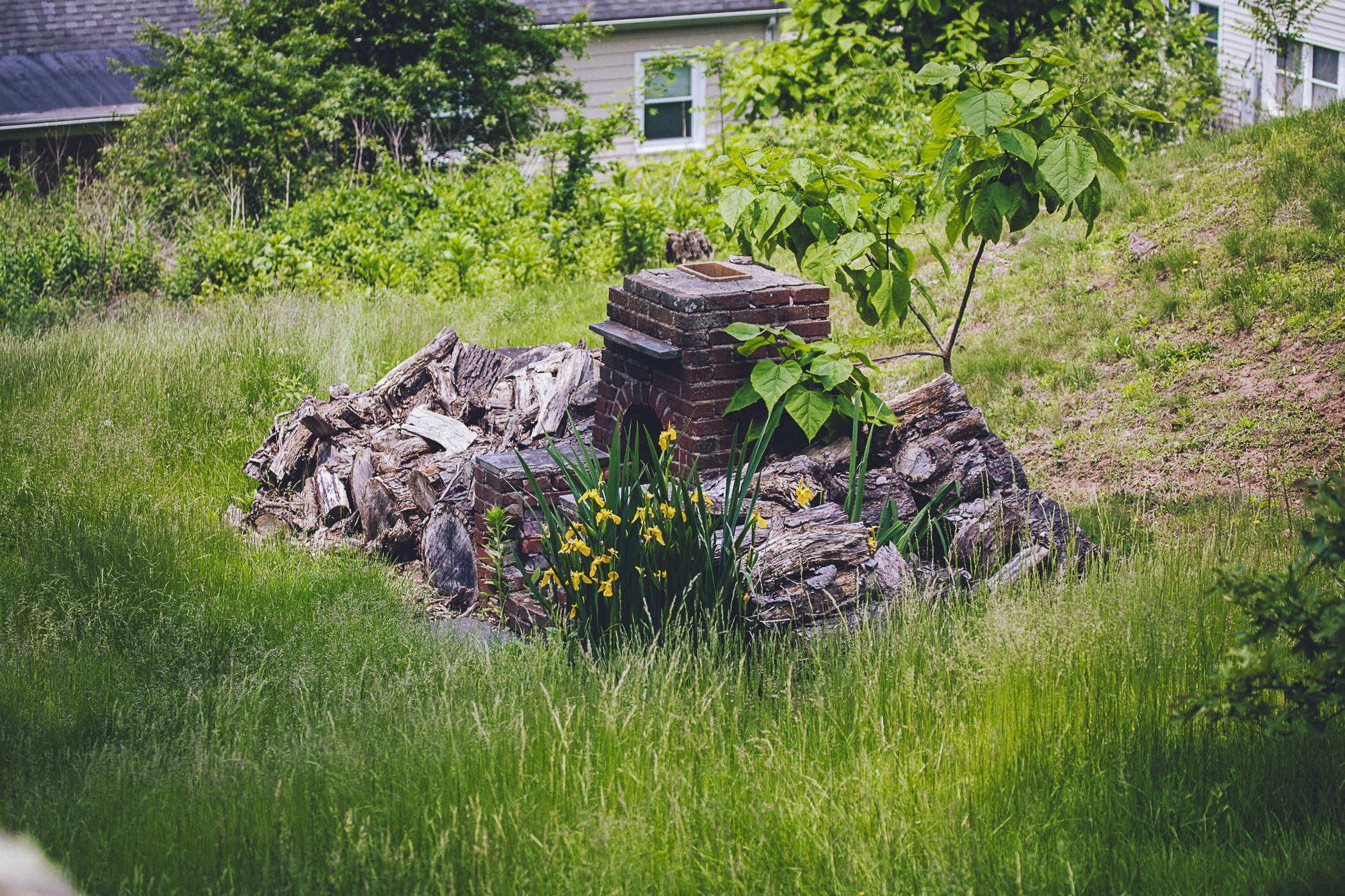 photo of a brick chimney structure surrounded by wooden logs, overgrown with tall grass and large flowers